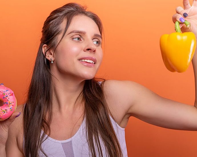 Woman in sports bra juggling choice between a donut and bell pepper on orange background.