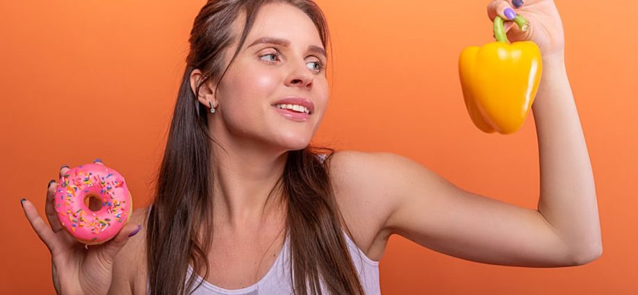 Woman in sports bra juggling choice between a donut and bell pepper on orange background.